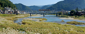 青空の下、山々に囲まれた自然豊かな河川敷で、橋の上を電車が走っているのが遠くに見える写真