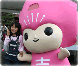 屋外で、頭と体がピンク色で目が黒丸のキャラクターと並んで、女の子が立ちながらピースサインをしている写真