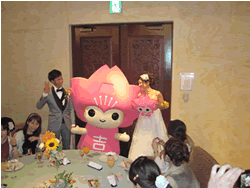 室内で、ピンク色のキャラクターを中央にして、タキシード服姿の男性とウェディングドレス姿の女性が横並びになり、着席している数人に見守られている写真