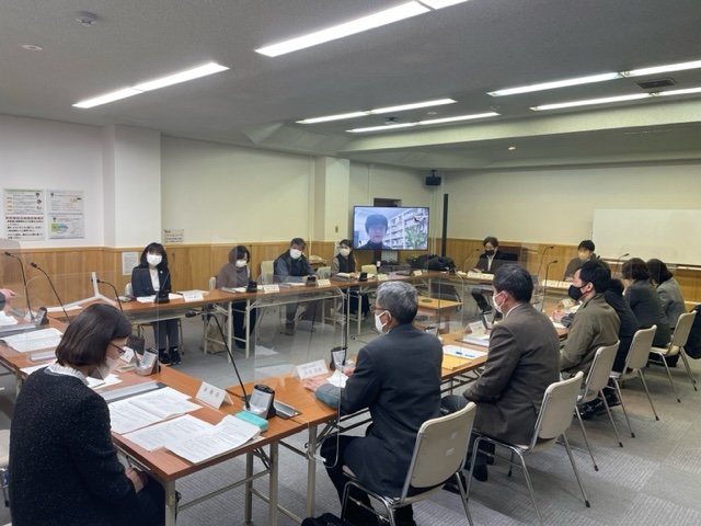 会議室に四角い形に机を囲むように配置し、スーツ姿の男性や女性が椅子に座り、資料を見ながら会議している様子の写真