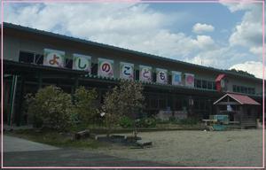 「よしのこども園」とカラフルな文字で書かれたパネルが設置されている幼稚園の建物外観の写真