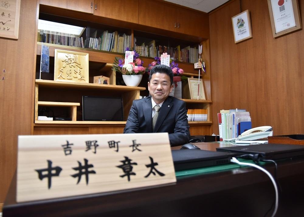 沢山の記念品や額縁に入った書類で飾られた室内で、「吉野町長 中井章太」と書かれた立て札が置かれているテーブルの上に肘を下ろし、座っているスーツ姿の男性の写真