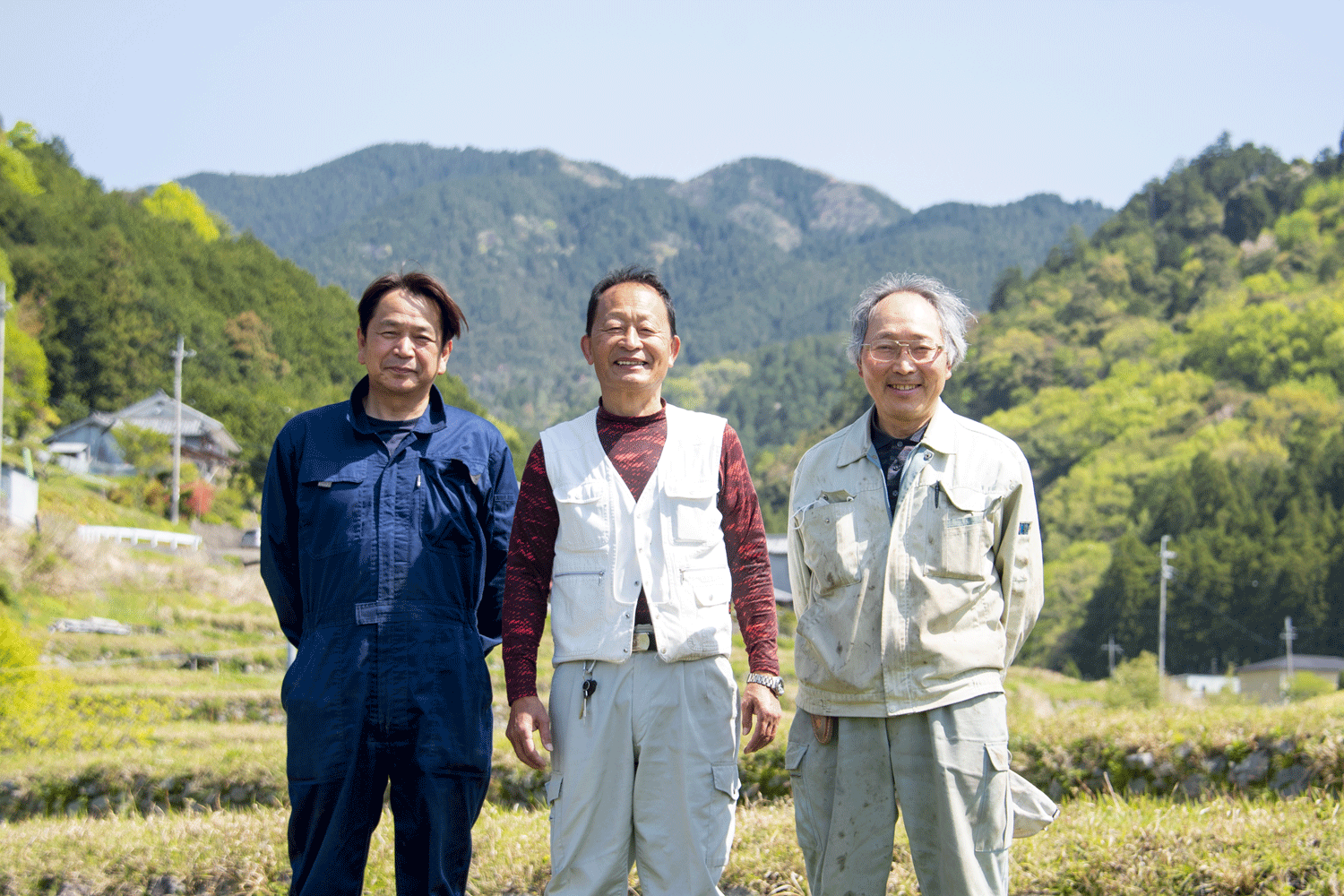 吉野正宗の酒米を作っている生産者3人が畑の前で笑顔でたっている。背景には龍門岳と青空が広がっている。