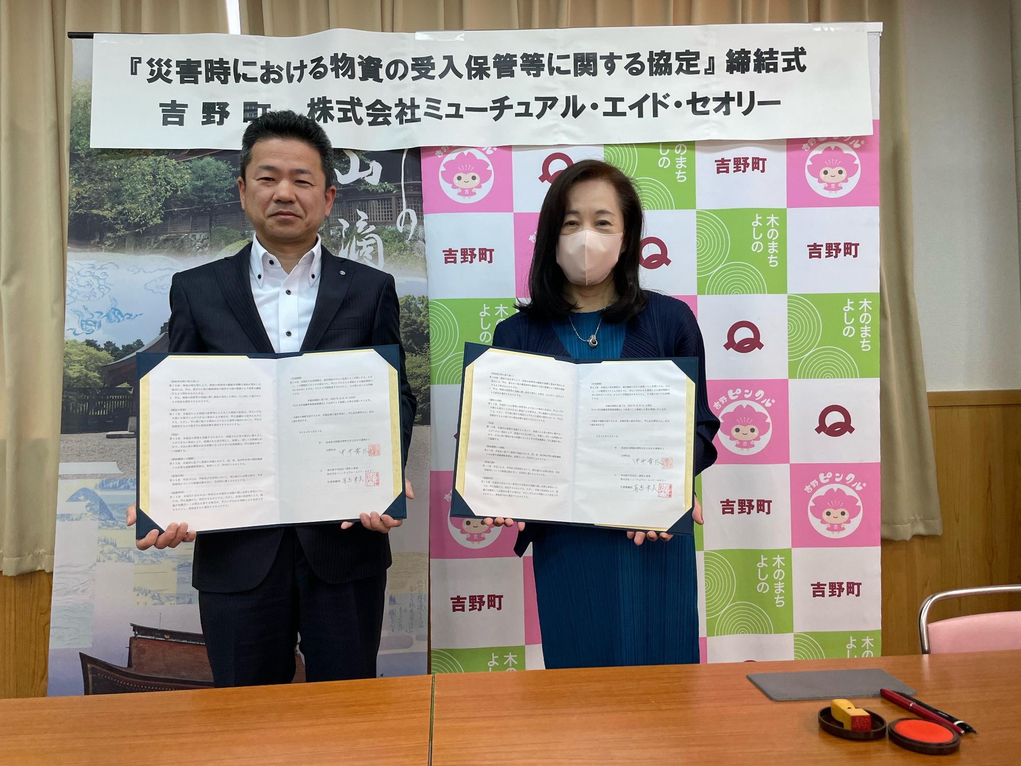 『「[災害時における物資の受入保管等に関する協定」締結式 吉野町 株式会社ミューチュアル・エイド・セオリー』と書かれた横断幕の前に書状を持った男性と女性の写真