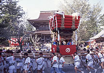 白いハッピを着た人達が神輿を担ぎ神社の前を通り過ぎている写真