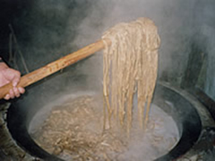 大きな釜の中で煮たれられた麺状の物体を、棒ですくい上げている写真