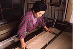 室内で、一人の女性が茶色の液体を木枠に入れて和紙を漉いている写真