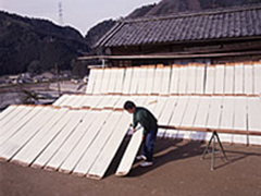 屋外で、一人の男性が、板に貼られた白い物体を天日干ししている写真