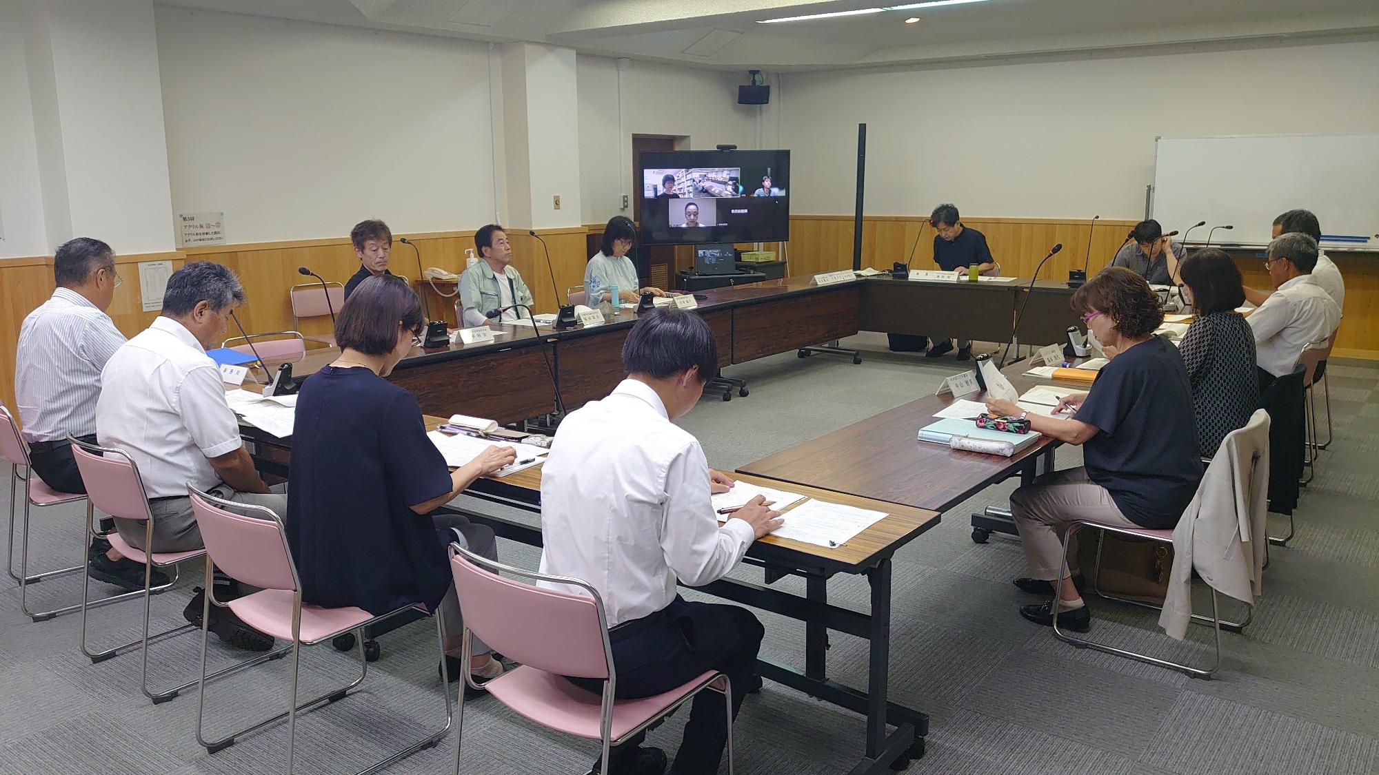 会議室で四角く囲むように配置された机にスーツ姿の男性や女性が座り、資料を見ながら会議をしている写真