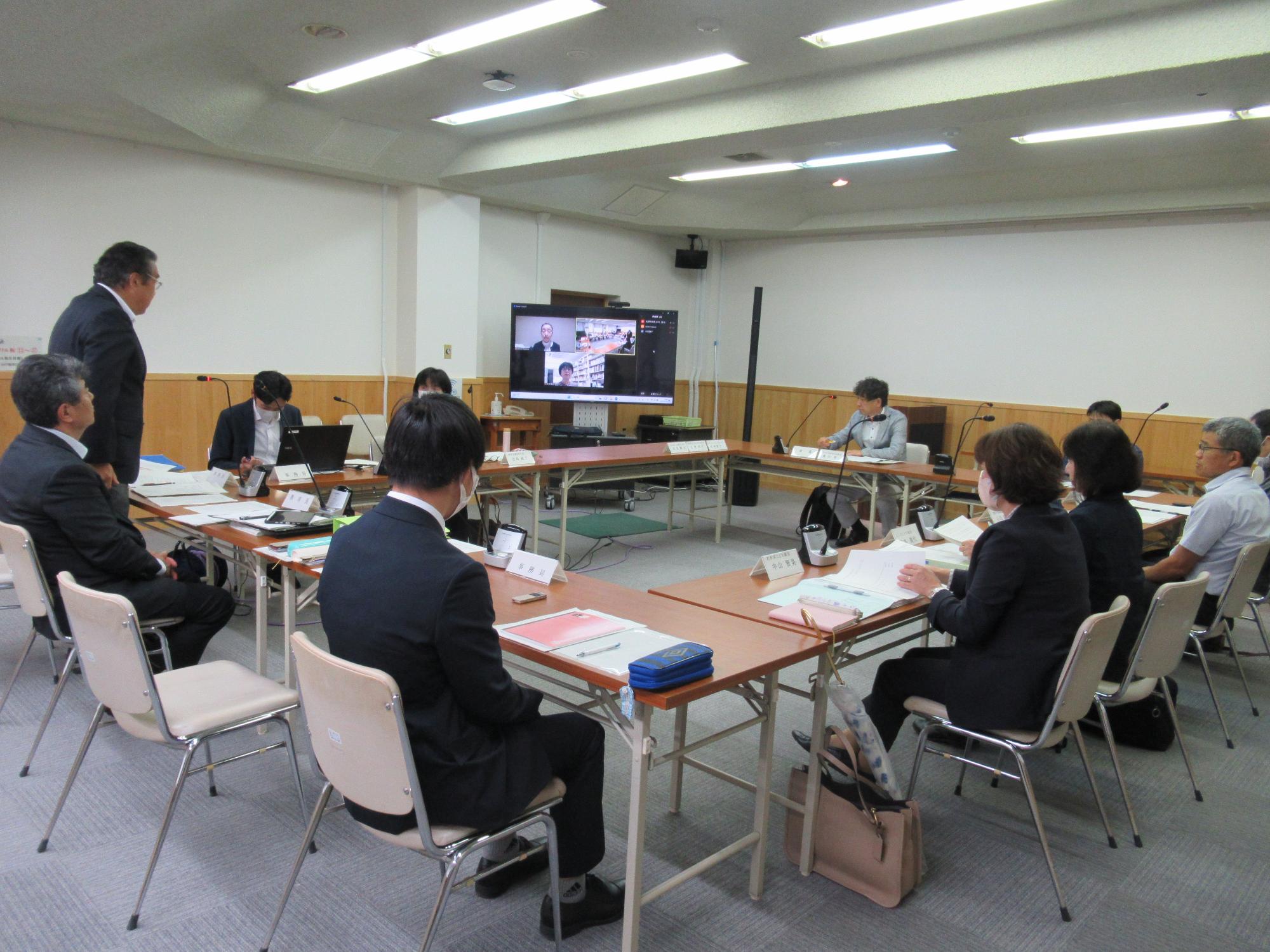 会議室で机を四角く配置し、スーツ姿の男性や女性が椅子に座り、正面のモニターに映し出された人とリモートで会議を行っている様子の写真