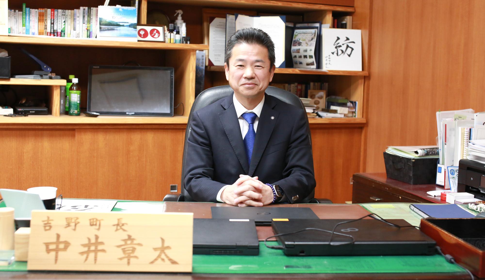 沢山の記念品や額縁に入った書類で飾られた室内で、「吉野町長 中井章太」と書かれた立て札が置かれているテーブルの上に肘を下ろし、座っているスーツ姿の男性の写真