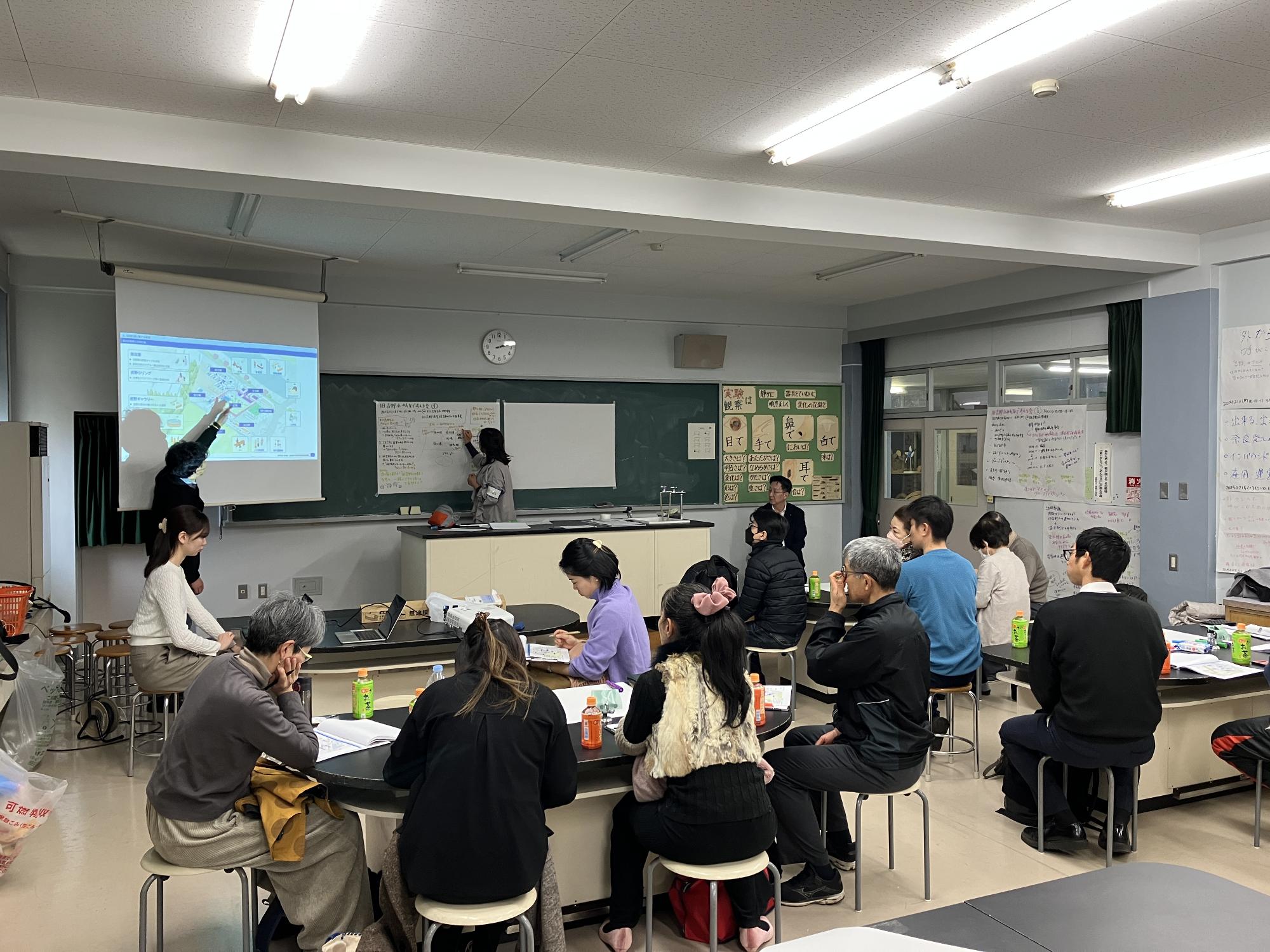 旧吉野小学校跡地で行われた旧吉小みんなで考える会の様子。プロジェクターには地図が表示され、黒板に模造紙を貼り説明内容を書き出している。説明を行っている人3名、説明を聞いている人約10名程度が椅子に座って資料やプロジェクター、黒板を見ている。