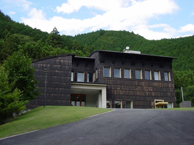 緑濃い山や木々に囲まれ、黒茶色の壁面が印象的な2階建て資料館の写真
